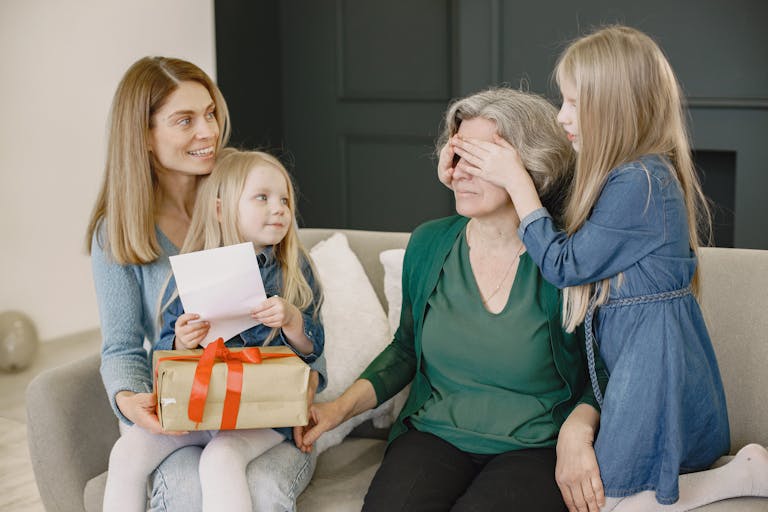 Grandmother receives a surprise gift from family, indoors with smiling relatives and joyful moments.