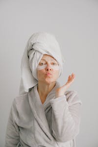 Woman with a towel on her head and eye masks, blowing a kiss in a robe.